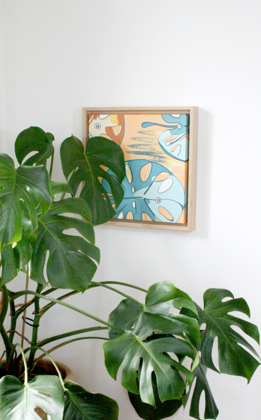 Monstera väggkonst nordisk tavla 30×30 - inramad tavla på vit vägg bredvid gröna monstera-växter i hem