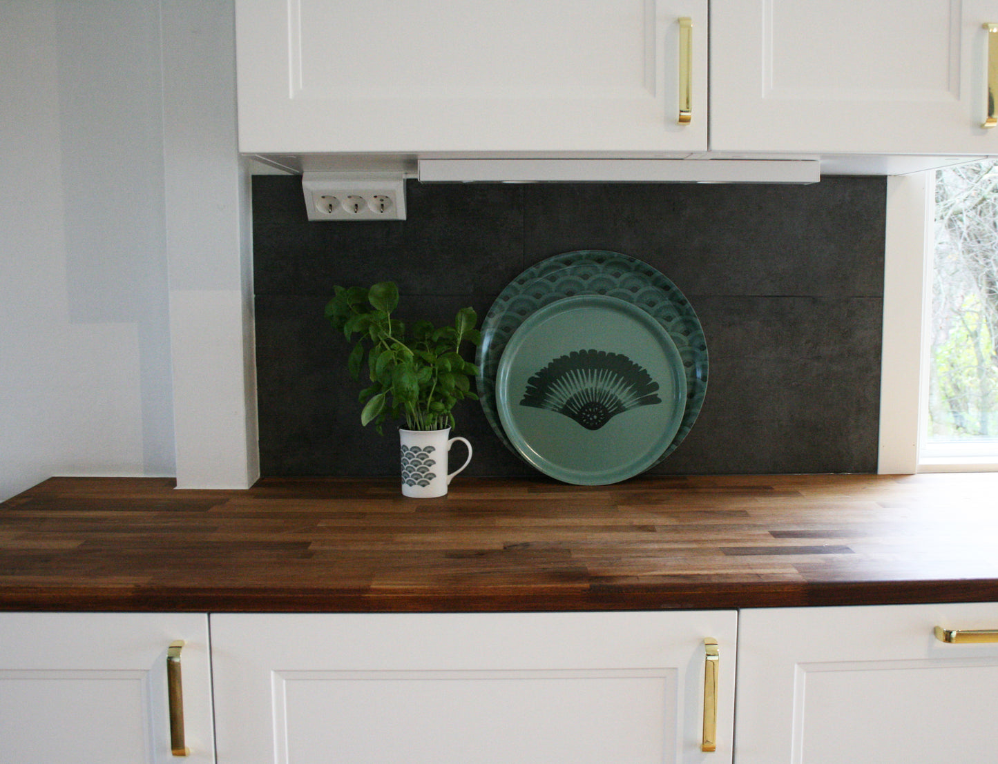 Serveringsbricka Solfjäder Björkfanér Skogsgrön 31cm, miljöbild. Bricka på bänk i kök mot antracit bakgrund och mugg med basilika.